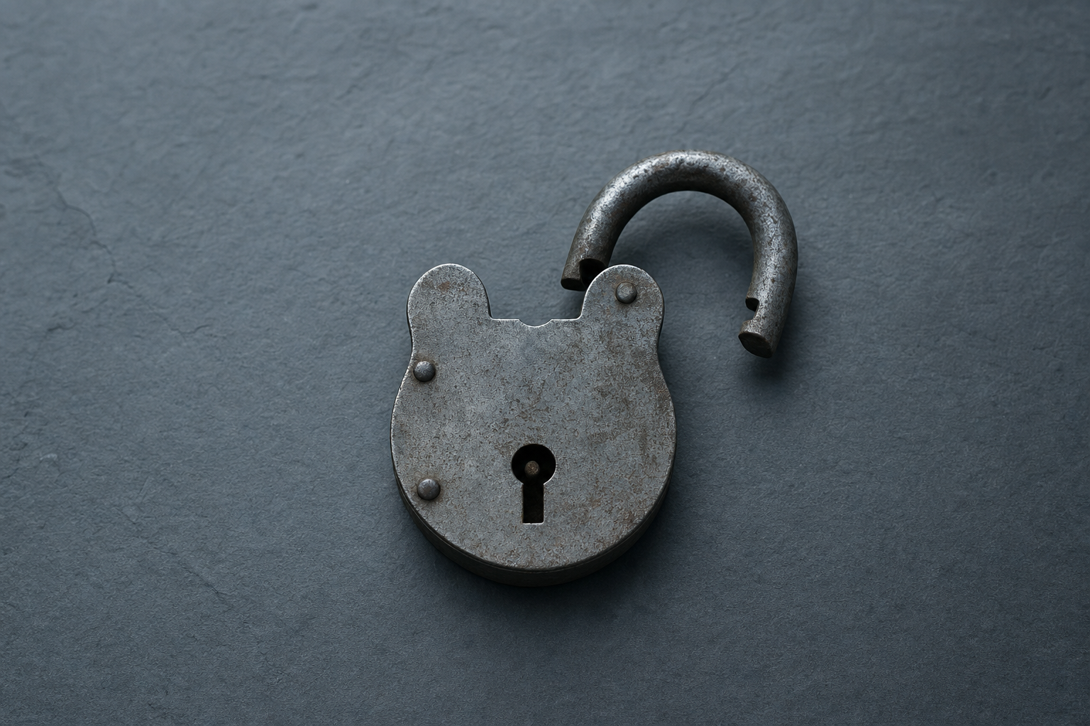 An aged steel padlock lying open on dark slate, its shackle swung clear of the body
